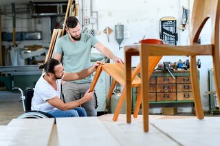 Zwei Handwerker arbeiten in einer Holzwerkstatt. Ein Mann im Rollstuhl inspiziert einen Tisch, während der andere ihn unterstützt.